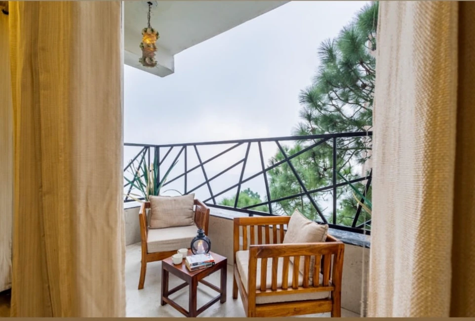 Hotel room balcony with wooden chairs, coffee table, soft curtains, and scenic outdoor views surrounded by greenery