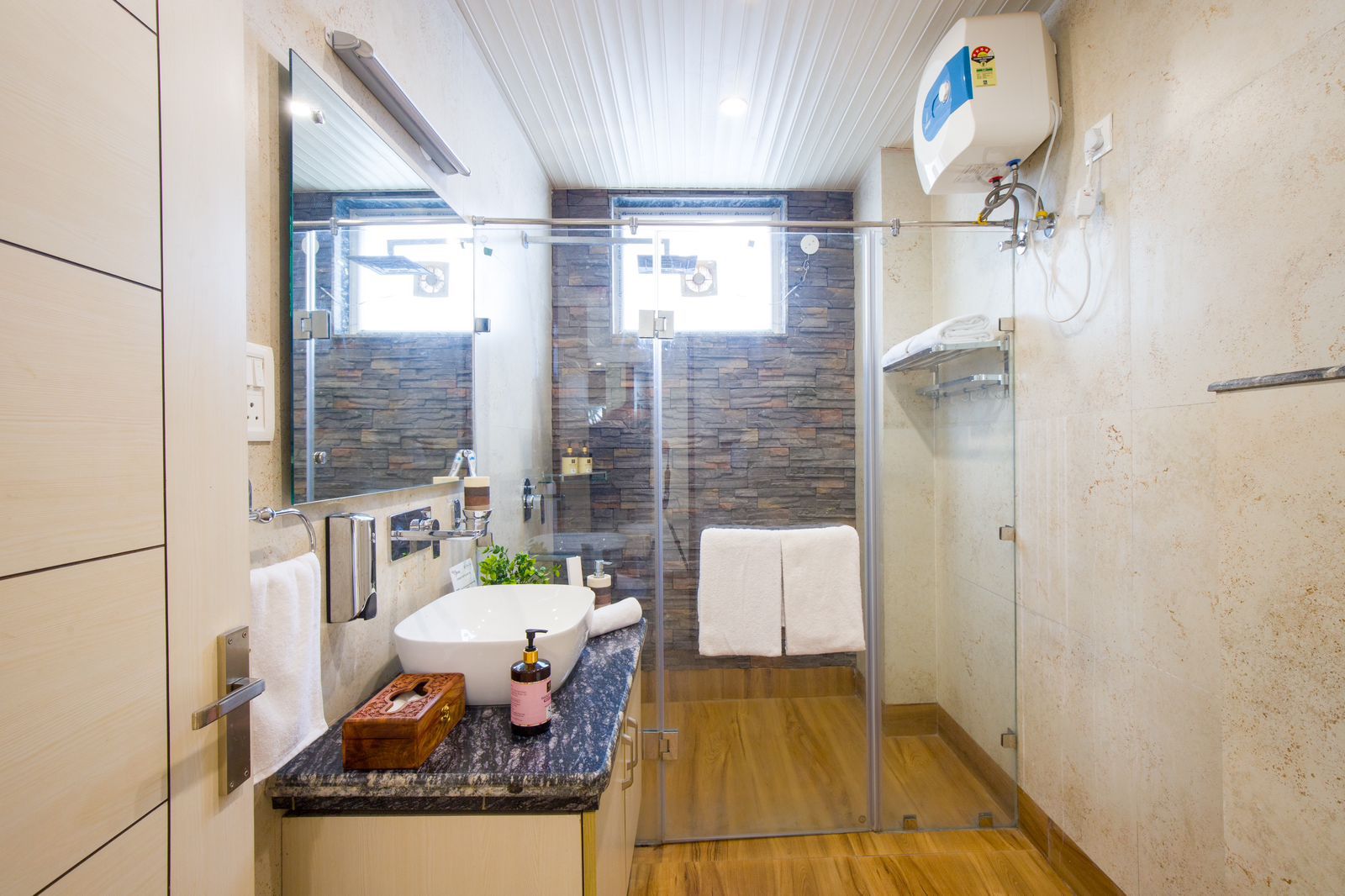 Modern hotel bathroom with glass shower enclosure, stone-textured walls, vanity mirror, fresh towels, and premium toiletries