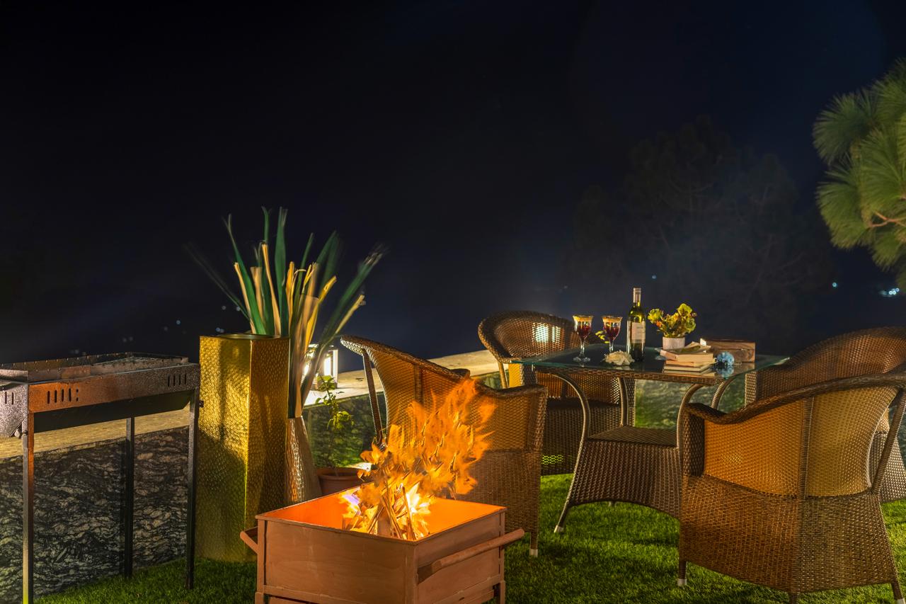 Outdoor hotel seating area with bonfire, wicker chairs, ambient lighting, and nighttime garden setting